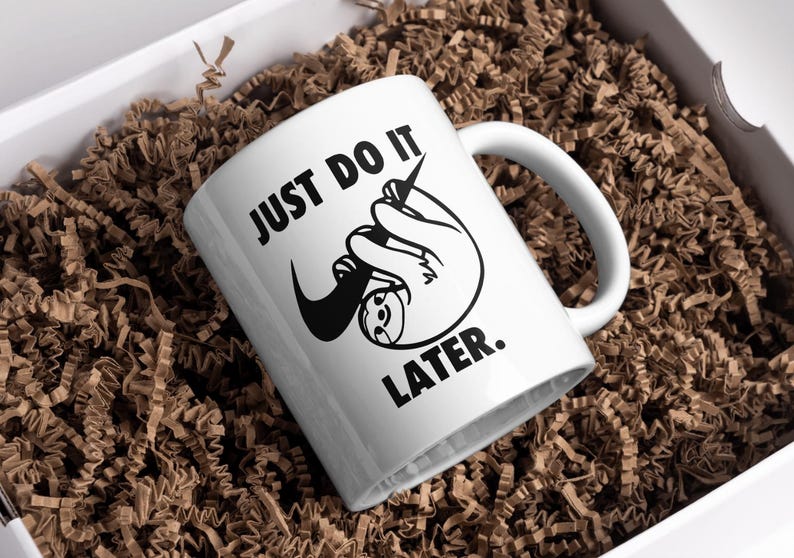 May include: White ceramic mug with a black sloth graphic and the text "JUST DO IT. LATER." The mug is inside a white box filled with brown crinkle paper, ideal for gifting.