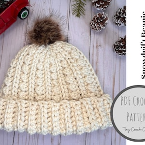 May include: Cream-colored, chunky knit beanie with a ribbed brim and a textured pattern. The hat is topped with a brown faux fur pom-pom. Text on the image reads "Snowdrift Beanie (Super Chunky Hat!)" and "PDF Crochet Pattern".