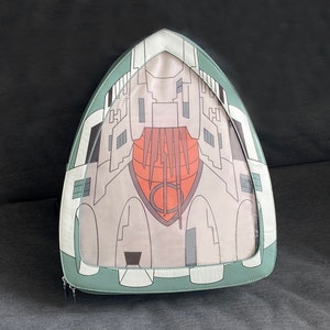 May include: A green and white backpack featuring a graphic of a spaceship on the front. The spaceship is grey and has a red and orange interior.