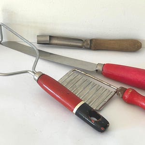 May include: A selection of vintage kitchen tools, including a potato masher, a knife, a crinkle cutter, and a peeler. The utensils have wooden and red handles, and stainless steel blades. The items are arranged on a white surface.