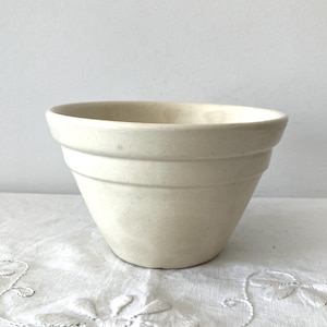 May include: A cream-coloured ceramic mixing bowl with a wide, open top and a tapered base. The exterior features two distinct horizontal ridges encircling the upper portion of the bowl, providing a textured grip and a classic, functional design.