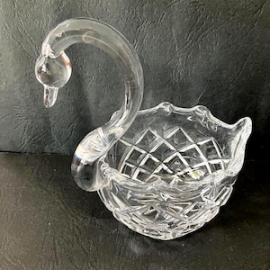 Op de afbeelding: Een heldere, zwanenvormige glazen schaal met een diamantgeslepen patroon. De nek van de zwaan buigt sierlijk en vormt een handvat, en de kom heeft een geschulpte rand. Het glas is transparant en staat op een donkere ondergrond.