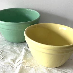 Op de afbeelding: Twee keramische mengkommen, één lichtgroen en één lichtgeel. Beide hebben een afgeronde vorm met een licht uitlopende rand. Ze staan op een wit, geborduurd doek. Geschikt voor bakken.