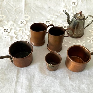 May include: An assortment of antique copper kitchenware, including small pots with long handles, mugs, and a small silver teapot. The copper pieces have a warm, reddish-brown hue, and the teapot has a tarnished silver finish. The items are arranged on a white, patterned surface.