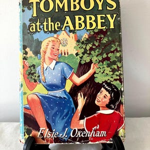 Puede incluir: Libro vintage titulado "TOMBOYS at the ABBEY" de Elsie J. Oxenham. La portada presenta una ilustración de dos chicas cerca de un árbol, con un edificio al fondo. El libro se muestra sobre un soporte negro.