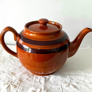 May include: A brown ceramic teapot with a rounded body, a curved handle, and a spout. The teapot has dark brown stripes around the middle. The lid has a small knob. The teapot sits on a white surface.