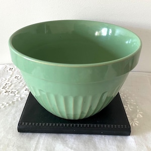 May include: A pale green ceramic mixing bowl with a ribbed exterior. The bowl sits on a black square stand. The bowl has a smooth, glossy finish and a rounded shape, ideal for baking and cooking.