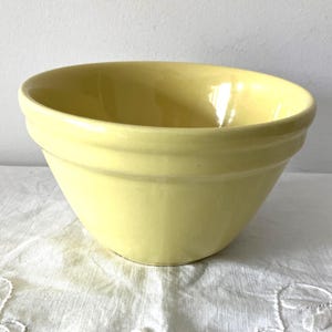 May include: A pale yellow ceramic mixing bowl. The bowl has a rounded shape with a wide rim and a slightly flared base. The exterior has a smooth, glossy finish. The bowl is sitting on a white, textured surface.