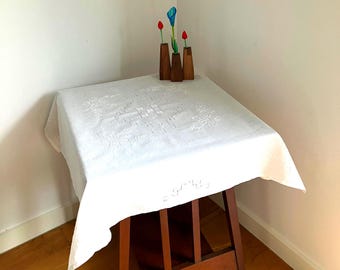 Könnte beinhalten: Eine quadratische weiße Tischdecke mit aufwendiger weißer Stickerei, die florale Motive und geometrische Muster zeigt. Das Tuch liegt auf einem dunkelbraunen Holztisch. Drei kleine Holzvasen mit roten und blauen Kunstblumen stehen darauf.