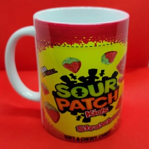 May include: White ceramic mug with a red and yellow design featuring the Sour Patch Kids logo and a strawberry candy. The text on the mug reads "Sour Patch Kids Strawberry Soft & Chewy Candy."