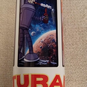 May include: White tumbler featuring a cartoon image of Bender from Futurama, holding a bottle and smoking. The background shows a space scene with planets and stars. The word "FUTURA" is printed in red.