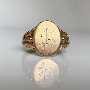 May include: A gold signet ring featuring an oval face with an engraved letter D in a gothic script style. The ring band displays intricate decorative scrollwork on both sides, providing a textured appearance to the metal surface.