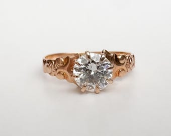 May include: A rose gold ring with a large, clear, round-cut diamond. The band features an ornate design with circular and curved patterns. The ring is set against a plain white background, highlighting the jewelry's details.