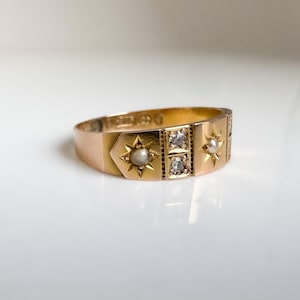 May include: A gold ring with star and gemstone details. The ring features a central section with two square-cut gemstones and is flanked by star-shaped designs, each with a small pearl. The ring is set against a white background.