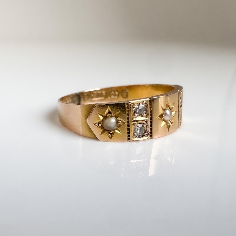 May include: A gold ring with star and gemstone details. The ring features a central section with two square-cut gemstones and is flanked by star-shaped designs, each with a small pearl. The ring is set against a white background.