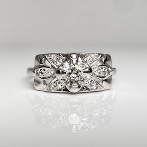 May include: A silver-toned ring featuring a wide, rectangular face with a central round stone surrounded by smaller, clear, faceted stones arranged in a geometric, floral-inspired pattern. The metal band is polished and reflective.