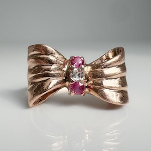 May include: A rose gold ring shaped like a bow, featuring a central vertical row of three round-cut gemstones. The top and bottom stones are vibrant pink, while the center stone is clear and colorless, set against the metallic bow design.