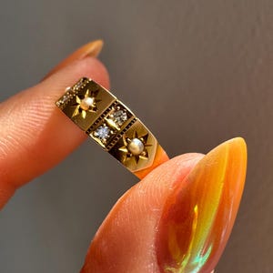May include: A gold ring with a rectangular design. The ring features star-shaped accents, small pearls, and clear gemstones. The ring is held between fingers, showcasing its intricate details and vintage style.