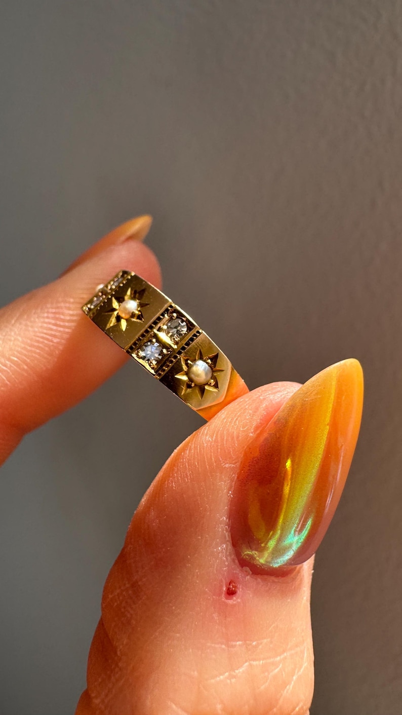 May include: A gold ring with a rectangular design. The ring features star-shaped accents, small pearls, and clear gemstones. The ring is held between fingers, showcasing its intricate details and vintage style.