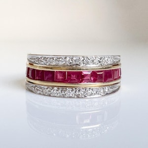 May include: A multi-band ring featuring a central row of square-cut red rubies set in yellow gold, flanked by two outer bands of silver-toned metal encrusted with small, clear, round-cut stones, creating a layered, wide-band jewelry design.