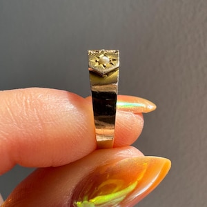 May include: A gold ring with a rectangular face, featuring a small pearl set within a starburst design. The ring's band is a simple, polished gold. The ring is held up against a gray background.