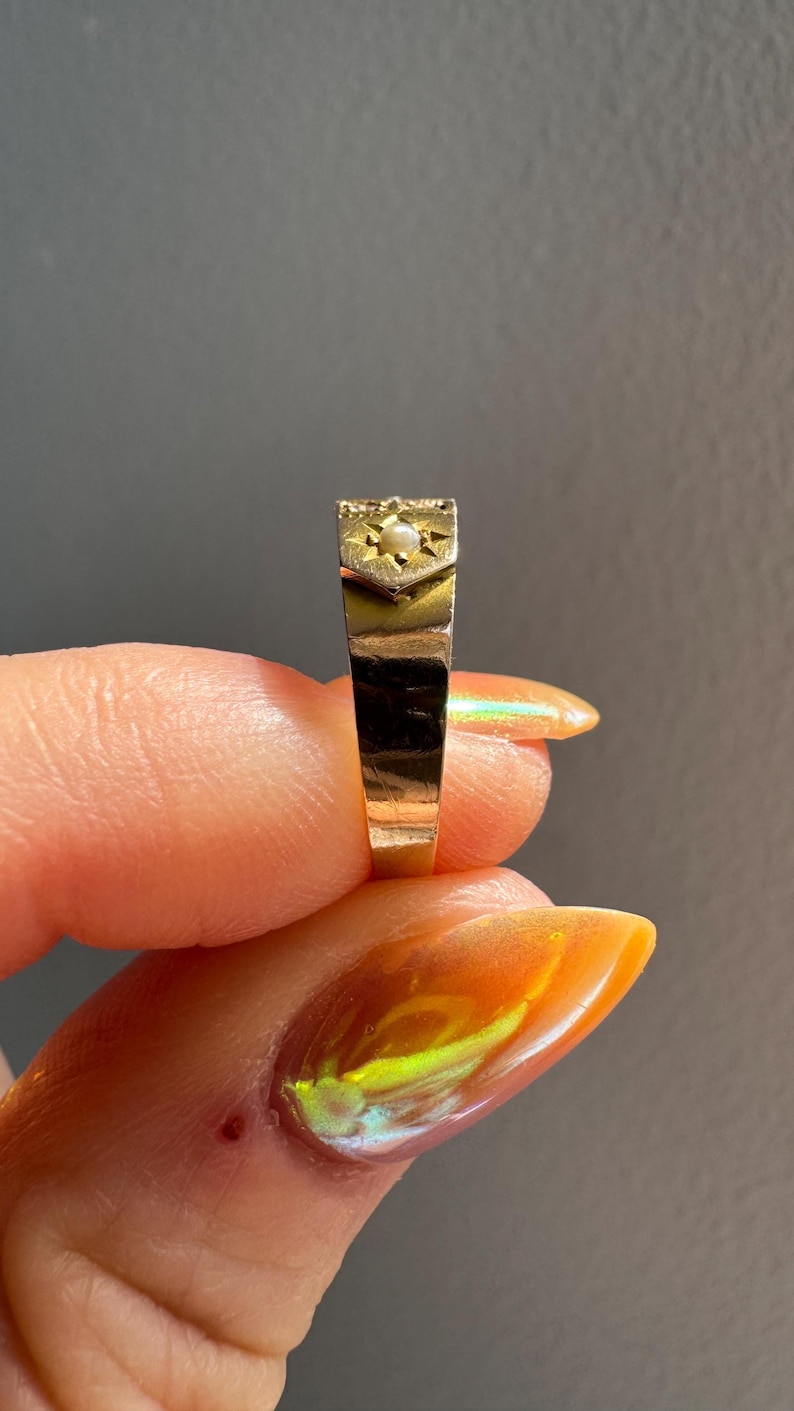 May include: A gold ring with a rectangular face, featuring a small pearl set within a starburst design. The ring's band is a simple, polished gold. The ring is held up against a gray background.