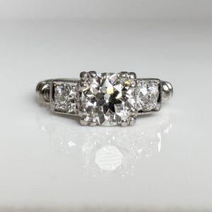 May include: A silver-toned metal ring featuring a large, square-cut central gemstone flanked by two smaller, round-cut stones. The setting is detailed with prongs securing each stone, and the band has a smooth, polished finish with a reflective surface.