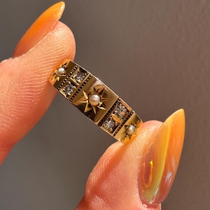 May include: A gold ring with a rectangular design, featuring star and square patterns. The ring is adorned with small pearls and clear gemstones. The ring is held between two fingers.