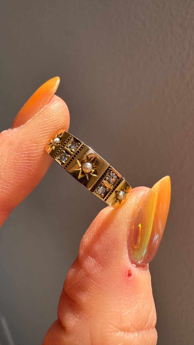 May include: A gold ring with a rectangular design, featuring star and square patterns. The ring is adorned with small pearls and clear gemstones. The ring is held between two fingers.