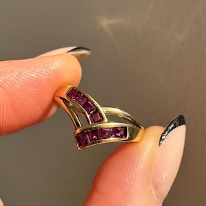 May include: A gold ring featuring a unique design with multiple rows of square-cut, deep red gemstones. The ring's band has a split design, creating an open, architectural look. The gemstones are set closely together, enhancing their rich color.