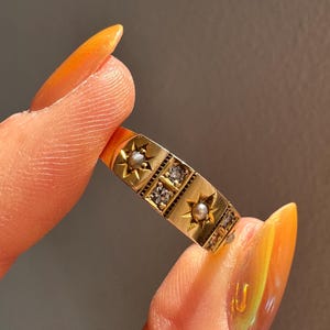 May include: A gold ring with a rectangular design featuring starbursts, small pearls, and clear gemstones. The ring is held between two fingers, showcasing its intricate details and vintage style.