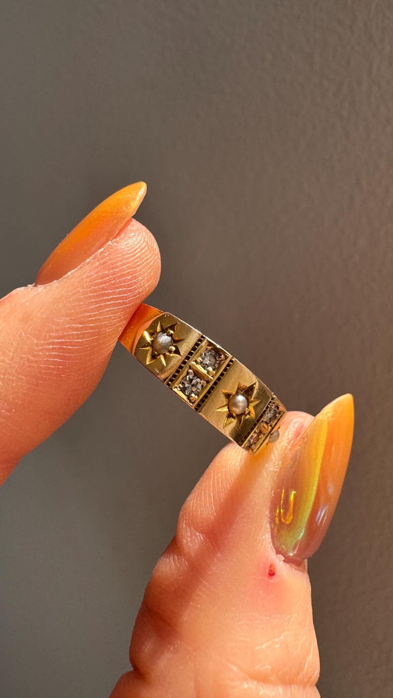 May include: A gold ring with a rectangular design featuring starbursts, small pearls, and clear gemstones. The ring is held between two fingers, showcasing its intricate details and vintage style.