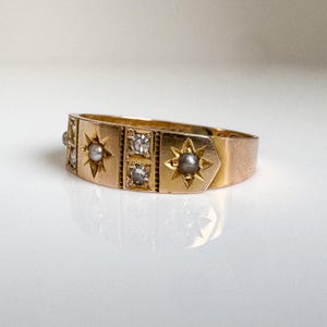 May include: A gold ring with a series of star and square designs. The ring features small, clear stones and gray pearls set within the star and square shapes. The ring is set on a white surface.