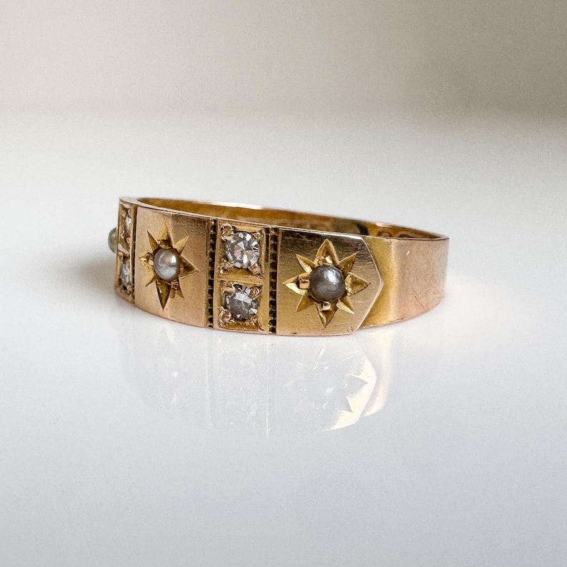 May include: A gold ring with a series of star and square designs. The ring features small, clear stones and gray pearls set within the star and square shapes. The ring is set on a white surface.