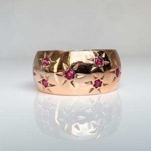 May include: A wide rose gold band ring featuring a polished finish and multiple star-shaped engravings. Each star setting contains a small, round, vibrant pink gemstone at its center, creating a repeating celestial pattern across the metal surface.