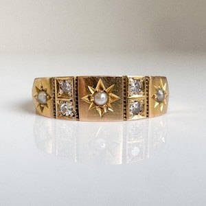 May include: A gold-toned ring with a repeating pattern of starbursts and square settings. The ring features a central starburst with a pearl, flanked by square settings with clear stones and smaller starbursts. The ring is displayed on a reflective surface.