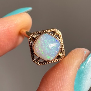 May include: A gold ring featuring a square-shaped opal gemstone. The opal has a milky white base with iridescent flashes of green and blue. The ring's setting has a decorative, openwork design.