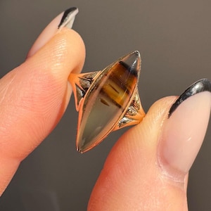 May include: A gold-toned ring featuring a large, elongated gemstone with brown and amber hues. The gemstone has a polished, smooth surface. The ring's band has intricate detailing on the sides, adding to its vintage appeal. The ring is held between fingers.