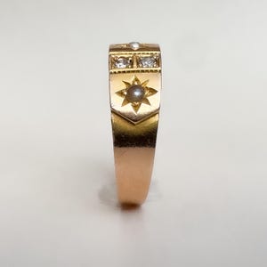 May include: A gold ring with a rectangular face featuring two clear stones and a pearl. A starburst design with a pearl is centered below the stones. A small pearl is set at the top. The ring has a polished finish.