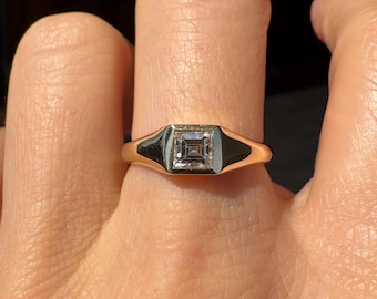 May include: A gold ring with a square, clear gemstone set in a bezel setting. The ring has a rectangular face with black enamel accents on either side of the gemstone. The ring is on a finger.