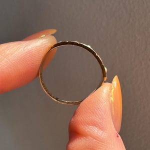 May include: A gold ring with a delicate design, held between fingers. The ring's band is thin, and the surface has small, evenly spaced details. The ring is held against a neutral gray background, highlighting its shape and craftsmanship.