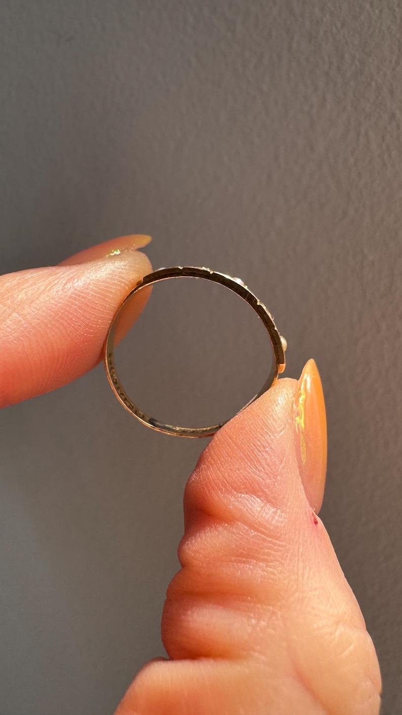 May include: A gold ring with a delicate design, held between fingers. The ring's band is thin, and the surface has small, evenly spaced details. The ring is held against a neutral gray background, highlighting its shape and craftsmanship.