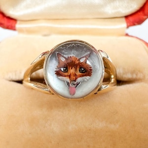 May include: A gold ring featuring a detailed, hand-painted fox portrait within a clear, circular setting. The fox has brown fur, black eyes, and a pink tongue. The ring is displayed in a velvet jewelry box.