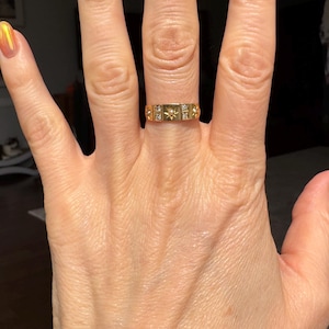 May include: A gold ring with a rectangular design featuring small starburst and square accents. The ring is worn on a hand with polished nails in shades of orange and yellow.