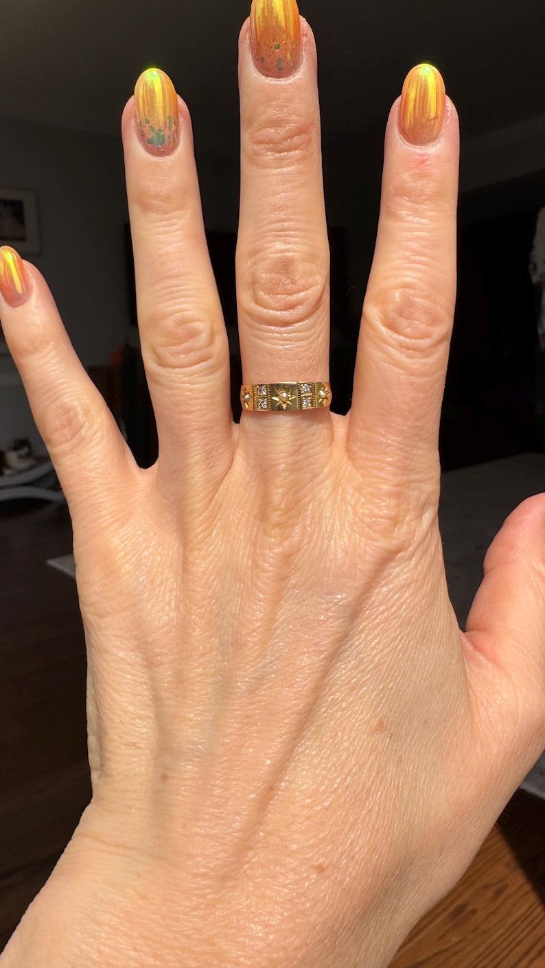 May include: A gold ring with a rectangular design featuring small starburst and square accents. The ring is worn on a hand with polished nails in shades of orange and yellow.