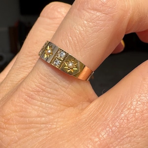 May include: A two-tone gold ring with a rectangular design. The ring features starburst and square accents, with small pearls and clear stones. The ring is on a finger.
