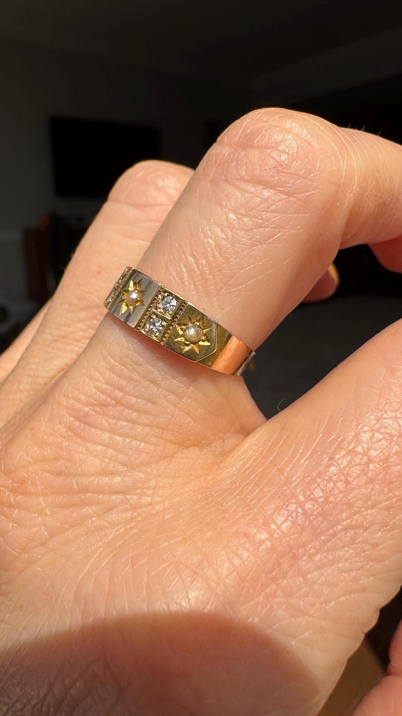 May include: A two-tone gold ring with a rectangular design. The ring features starburst and square accents, with small pearls and clear stones. The ring is on a finger.