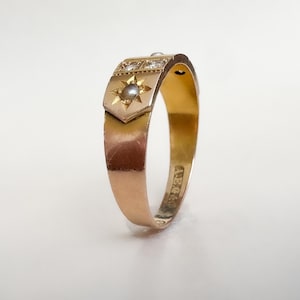 May include: A gold ring with a starburst design and a small pearl. The ring has a rectangular top with small diamonds and a starburst design with a pearl in the center. The band is smooth and polished.