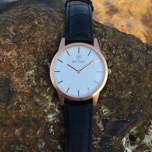 May include: A rose gold wristwatch with a white face and black leather strap. The watch face has a simple design with black hour and minute hands and a small logo that says "BATTUTA".