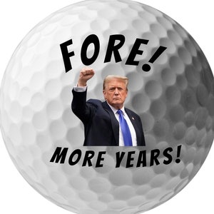 May include: A white golf ball with a picture of a man in a suit with his fist raised in the air. The text "FORE! MORE YEARS!" is printed on the ball.